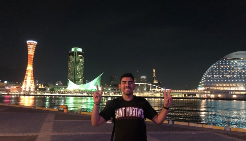 SMU student holding up peace signs in front of a lit city skyline along water at night
