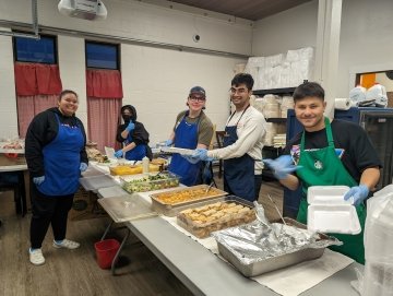 Students in aprons serve food in a line at community kitchen